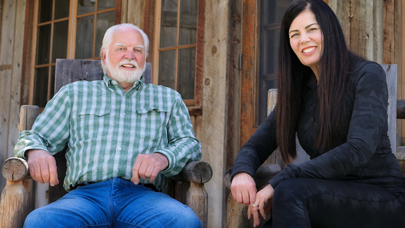 New Day Filmmakers Joe Phelps and Rosemary Smith sit side by side on wooden chairs, smiling at the camera. Joe wears a green and white plaid shirt and Rosemary is dressed in black.