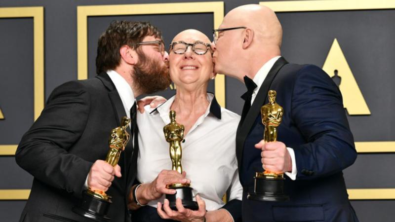 Julia Reichart, with a bald head and round glasses, holds an Oscar. She smiles with her eyes closed, and is kissed the men on either side of her, each holding an Oscar statue.