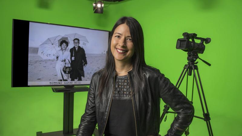 Kimi Takesue smiles for the camera in a studio with green-screen walls. Behind her, a video camera on a tripod and a monitor showing a black and white image of her grandparents on the beach with parasols.