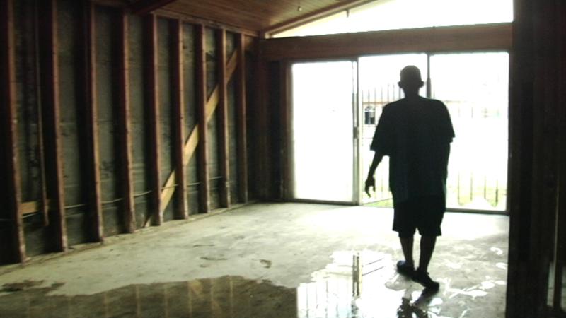 A man stands inside a building where the wall beams are fully exposed and water pools on the cement floor. He is silhouetted against bright windows in the background.