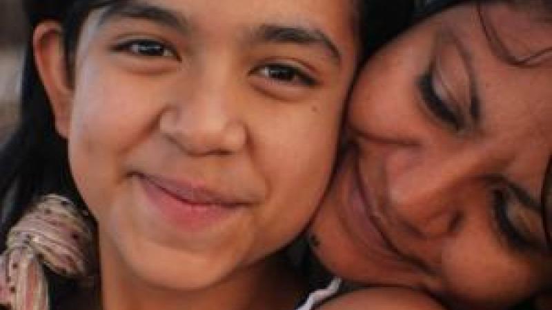 A still from the New Day film Life on the Line. A young Mexican-American girl, Kimberly Torrez smiles sweetly towards the camera. A woman, likely her mother Vanessa Torrez sits behind Kimberly and rests her head on her shoulder. She turns her face into Kimberly’s neck with a loving smile.