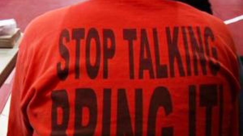 A still from the New Day film Making Noise In Silence. The back of a person sitting on a bleacher. They are wearing a bright orange t-shirt that reads “Stop Talking, Bring It!” in large black letters. The ground is a red gymnasium floor with a wrestling ring.