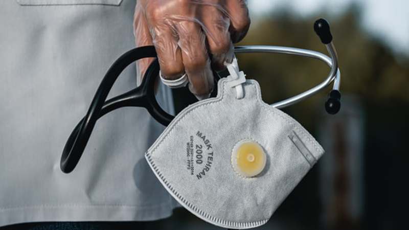 A latex-gloved hand holding a disposable respirator and a stethoscope. The hand is next to the torso of the person's white lab coat.