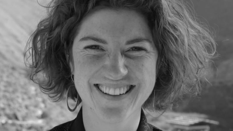 In this black and white photo, New Day filmmaker Meghan Shea looks directly at the camera and smiles widely. Her medium-length hair is above her shoulder and she wears hoop earrings.