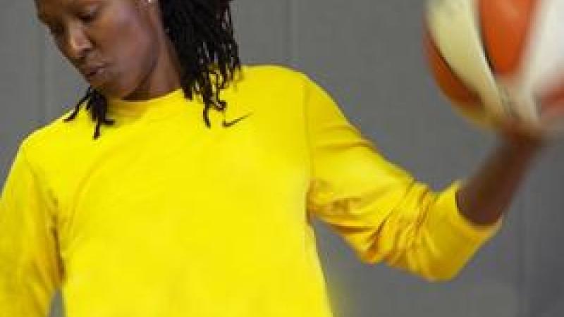 Chamique Holdsclaw, an African-American woman with her locs pulled back looks intently toward the ground, lips pursed. She wears a bright yellow Nike long-sleeved shirt and holds a basketball aloft in one hand.