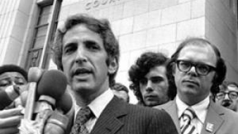 A black and white still image from the film. The camera looks up from below at Daniel Ellsberg, a white man in a pinstriped suit, who speaks into a mass of TV reporter microphones that are taped together. He stands in front of tall, pale building with “United States Court House” embossed on the exterior wall. He is surrounded by men of various ethnicities who listen on or jockey into position to get their microphone closer to him.