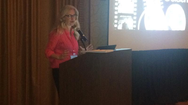 Laurel Chiten wears a bright pink blazer and stands in front of a podium on the stage at the New Day House in the National Media Market. There is a bright screen on her left with an image of film strips on it.