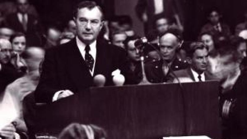 A black and white image from the film Nuremberg: Its Lesson for Today. A man in a suit stands at a podium with a serious expression looking slightly off to the side. Behind him sits a full audience of people, looking in different directions with serious expressions on their faces. In front of the podium sit women with headsets on and large stacks of paper in front of them.