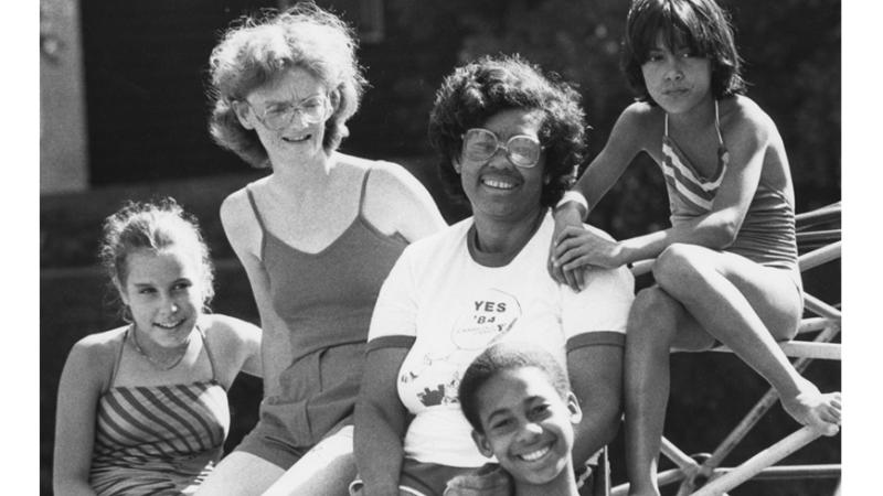 In this black and white photo from the 1980s, five teens and children of various races sit on playground equipment smiling on a bright summer’s day.