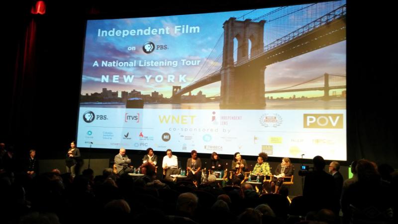 An auditorium with a large screen on the stage. On the screen is an image of Brooklyn Bridge and the words “Independent Film on PBS A National Listening Tour New York” in white text. On stage a panel of people of various races sit in a row, some leaning over in discussion with one another.