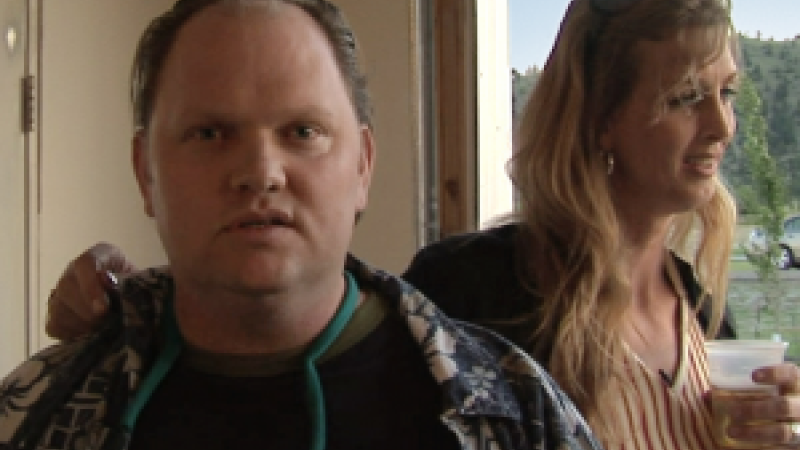 A still from the New Day film Prodigal Sons. A young man faces the camera with a slightly surprised expression as if he wasn’t expecting a picture to be taken. His sister, New Day filmmaker Kimberly Reed rests her hand on his shoulder. She is standing behind him and facing to the side with a drink in her hand. Behind her is a window with trees outside.