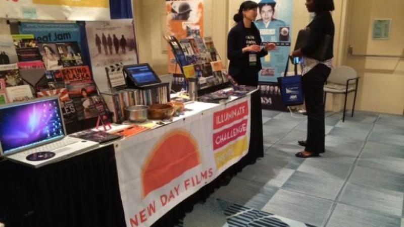New Day filmmaker Debbie Lum converses with an attendee at the New Day exhibition booth filled New Day films promotional materials and a laptop to screen trailers.