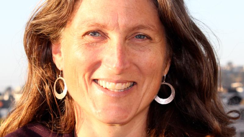 A headshot of New Day filmmaker Reagan Brashear. She smiles and looks into the camera with the sun shining across her face. She wears long earrings with silver circles at the bottom. In the background is a light-blue sky.