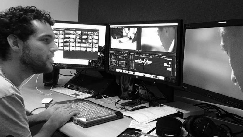 New Day Filmmaker Reid Davenport is sitting at a desk with his hands on a keyboard in a video editing room. He faces three video monitors and has a small smile on his face.