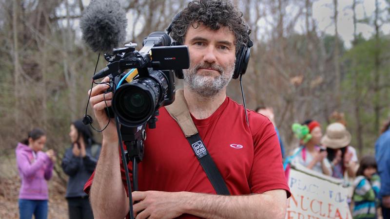 Robbie Leppzer on location filming at a protest holds his video camera and wear headphones.