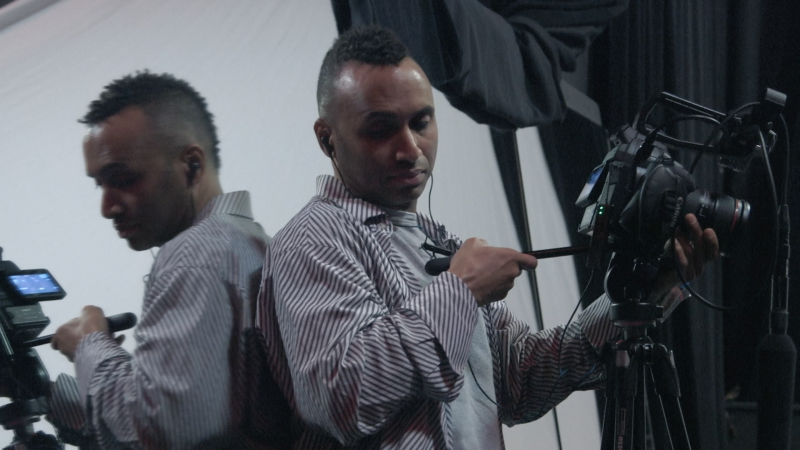 Rodney Evans, a Black man, focuses a video camera on a tripod while looking down into the camera's monitor. He leans against a mirrored wall, his face and the camera reflecting back.