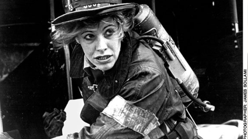 In this black and white still from the film, a female firefighter is fully geared up in a heavy coat, gloves and helmet as she races at the scene of a fire. She wears an oxygen tank on her back and looks out towards the distance, with determination on her face.