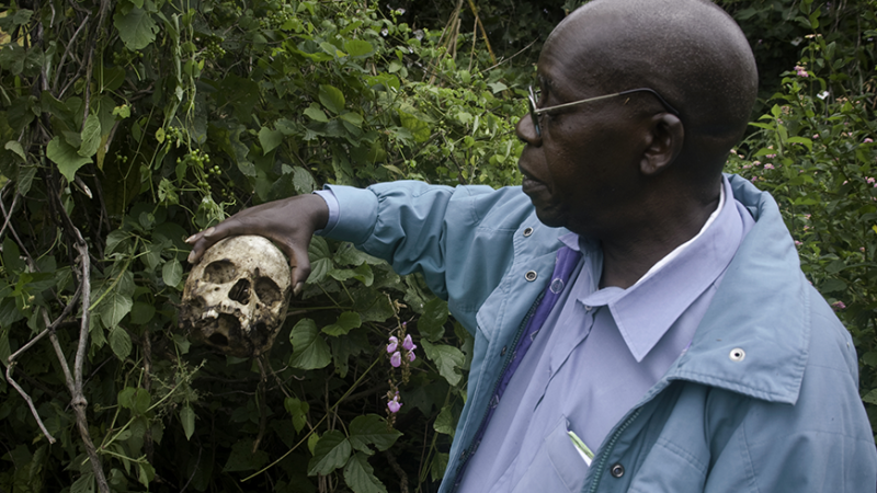 In this photo from the New Day Film The Reckoning: The Battle for the International Criminal Court, a dark skinned black man stands in a grove of leafy green bushes. In his right hand. He holds a human skull with its jaw missing.