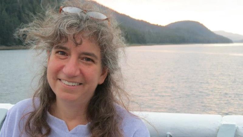 A headshot of New Day filmmaker Ellen Brodsky. Ellen smiles and stands on a boat with the ocean and mountains in the background. Her glasses are on top of her hair which is blowing in the breeze.