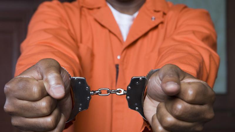 A Black man's arms are extended in front of him, hands in fists, cuffs around his wrists. He wears an orange prison jumpsuit.