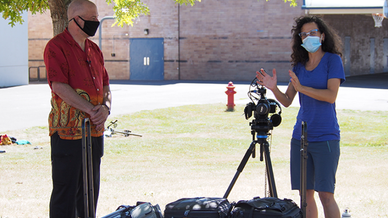 New Day Member Cheryl Green stands in a small patch of shade at an outdoor film shoot with Karl, the star of “TBI & My Longest Ride.” They are white, and both wear masks, standing at opposite ends of a cart full of video gear. Cheryl, by her camera and tripod, looks toward the distance and gestures as Karl watches her discuss the next shot.