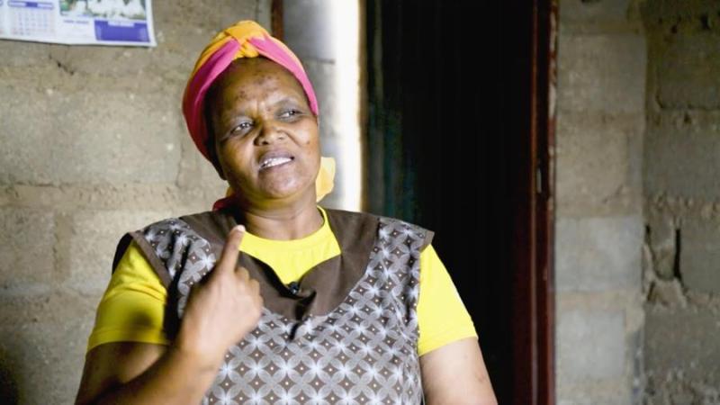 A Black woman wearing a yellow shirt with a brown patterned dress over it and a pink and orange head wrap. She sits in front of a cinder block wall with an open door. She is looking to the side and has her index finger raised.