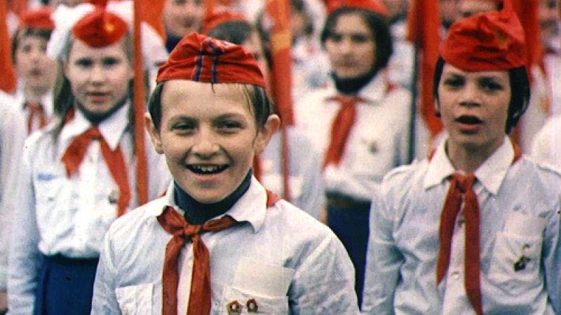 Russian Children stand on guard wearing military outfits which include red hats and red scarves, white shirts with pins on their pockets.