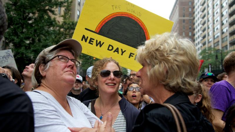 Three people stand in a circle at the People’s Climate March looking in different directions. A large yellow poster with the New Day Films Logo is hoisted behind the middle woman’s head. The poster has various writings on it, including the words “Save the World” written above the logo. People fill the street moving forward behind the three women and tall city buildings rise up on either side of the frame.