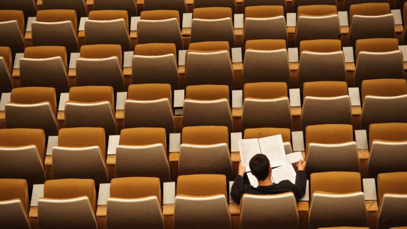 Overhead view of empty, orange seats in an auditorium style classroom. One student in the lower right sits and reads from two open notebooks.