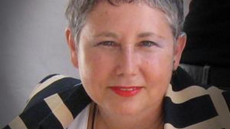 A headshot of New Day filmmaker Sandra Shulberg. A woman with short gray hair and a patterned blazer looks into the camera and smiles. The background is mostly gray with a spotlight behind her head and a stripe of black to the right.