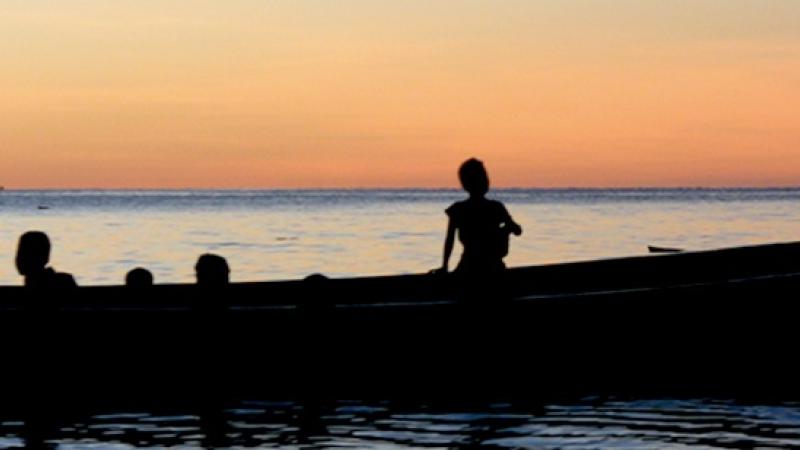 A still from the New Day film Sun Come Up. A silhouette of a large canoe in shiny, calm waters with a deep orange sunset behind. Several people’s heads at different levels show them standing or sitting in the boat. Two people on the right stand at the back of the boat with an oar. A few short buildings disrupt the mostly flat horizon.