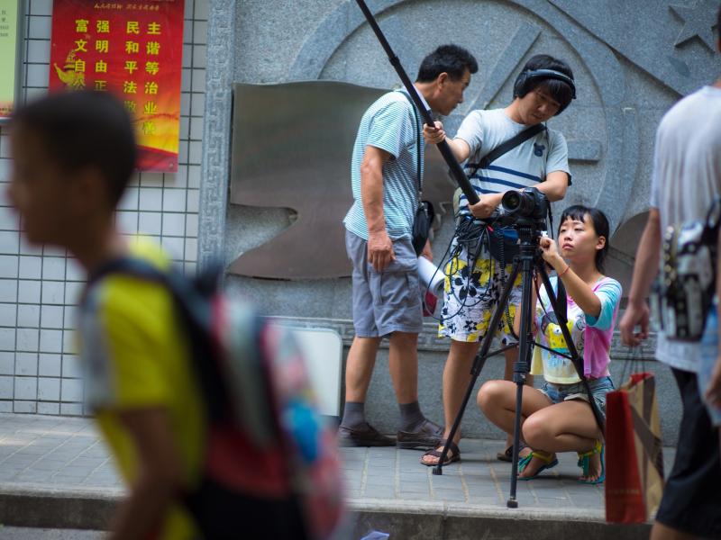 Yoyo behind camera with a sound person, outside of a school gate, in the foreground a young student and some parents walk by