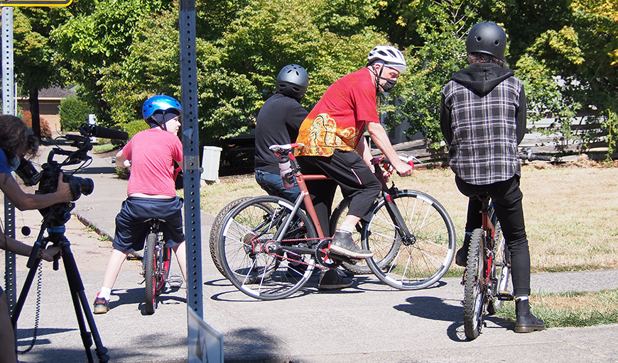 On the left edge of the frame, Cheryl, in a medical mask, bends over a camera mounted on a tripod and films Karl and his three sons as they wait to ride their bicycles. Karl also wears a mask. It is a summery day with sunshine and green trees in the background.