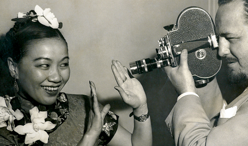 Chinese-American filmmaker Li-Ling Ai wears a silk patterned dress with flower blossoms pinned by the neckline and in her pulled-back hair. She smiles and playfully holds up her hand to create a frame around her face. White photograph Rey Scott wears a light linen suit and holds a film camera to his eye, the lens just an inch from her hands. The photograph is black and white.