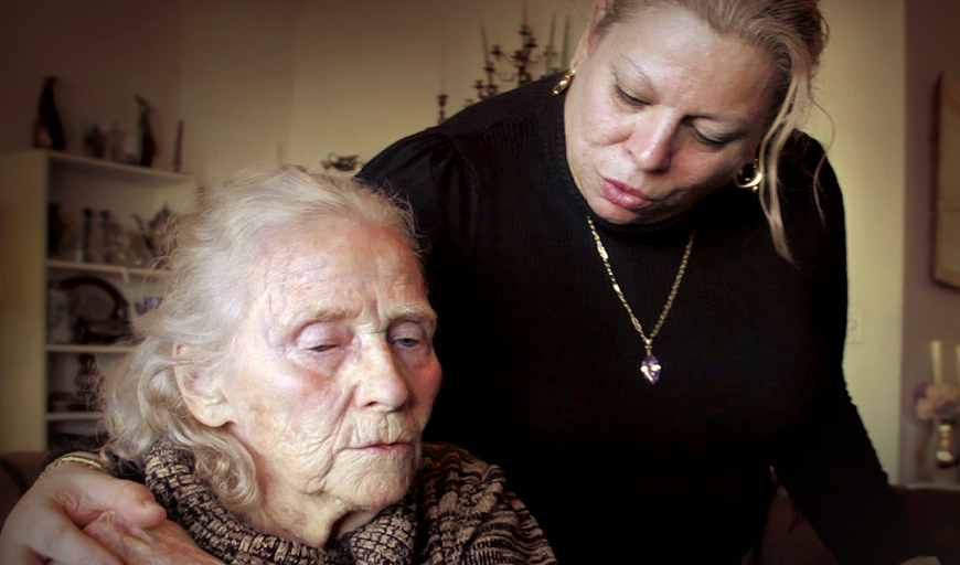 A middle age woman is standing over an elderly woman with her arm around her. 