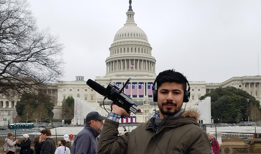 New Day Films director Ahmed Mansour is holding a film camera in one hand and looking straight at the camera. He has dark hair and a short beard. Behind him is the United States’s Capitol building.