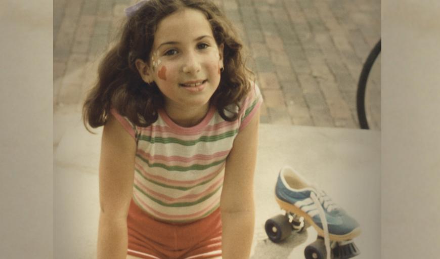 In this dated photo from the seventies, a young girl wearing a striped shirt and red shorts looks up at the camera and gently smiles. She has a heart and a flower painted on her cheek. To her right is an old-fashioned roller skate with thick, black wheels.