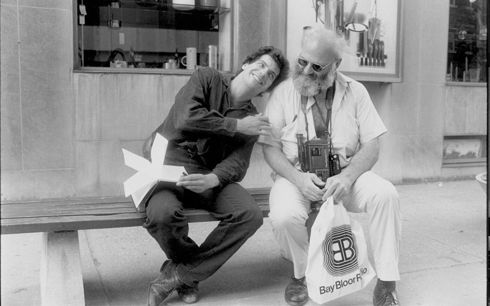 In this black and white photo, two men sit on a bench and smile at each other. On the left, actor Shane Fistell holds an open notebook on his lap and a pen in one hand. He lays his head on the shoulder of the famous physician Oliver Sacks. Dr. Sacks has a thick, white beard. He looks downward, wears a camera around his neck, and holds a white shopping bag.