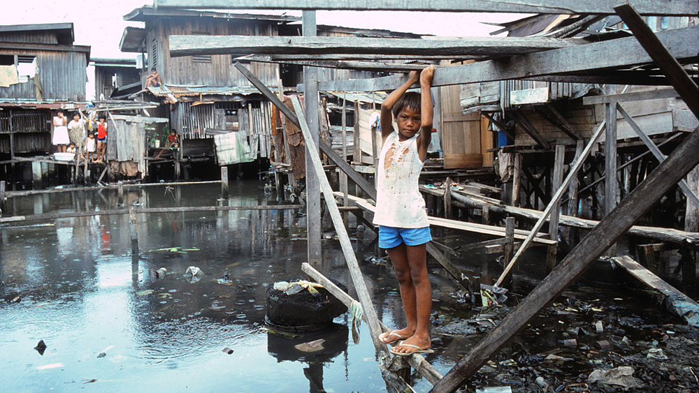 Outside, surrounded by wooden shacks, a young Asian boy stands above water, and holds onto a wooden plank above him and stares at the camera with a forlorn expression. He wears a ripped, white sleeveless t-shirt and blue shorts.
