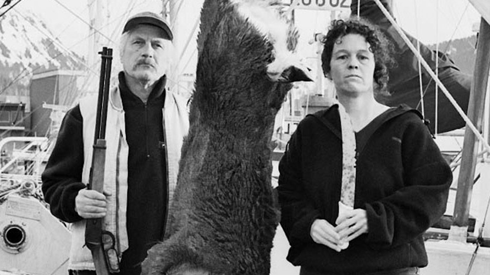 In this black and white photo, a woman and man stand on a dock with a dead, hanging deer between them. The man holds a hunting rifle. Behind them is a boat.