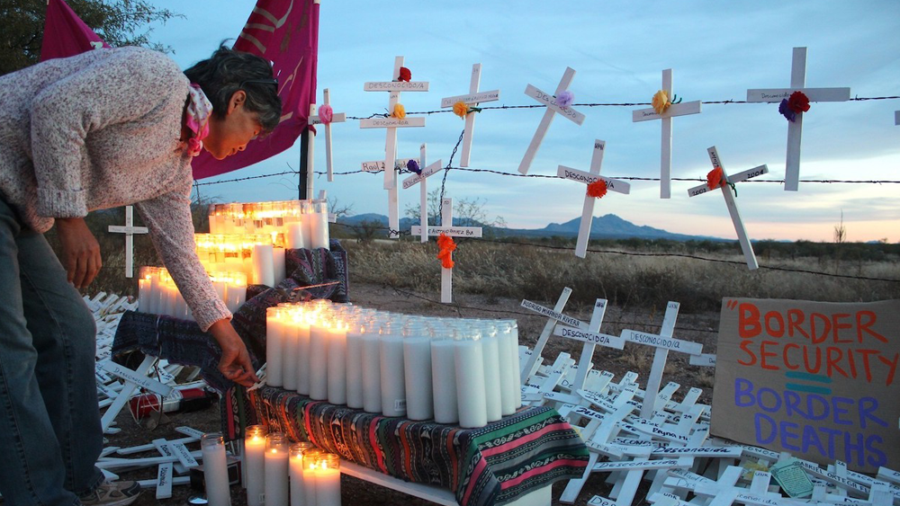 A still from the film “Undeterred” shows a memorial at a barbed wire fence on the Arizona border at dusk. A woman bends over a table full of memorial candles where some are lit. She’s surrounded by white crosses with the names of people who have died at the border written in black. Leaning against a barbed wire fence is a sign that reads “Border Security = Border Deaths.”