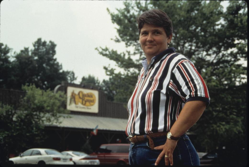 A white woman with short hair stands outside a Cracker Barrel restaurant and looks over her shoulder to smile toward the camera.