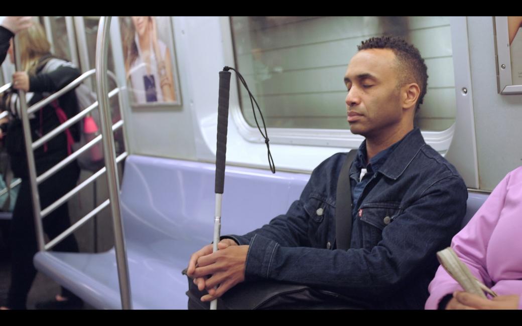 A still image from Vision Portraits shows Rodney Evans, a Black man, sitting on a subway. His eyes are closed, his face serene, and his fingers rest gently around his white cane.
