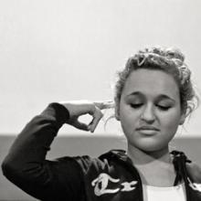 A black and white still from the New Day film Deaf Jam. Young ASL poet Aneta Brodski closes her eyes with a vulnerable but dignified expression on her face. She holds out one finger pointed out towards her ear. She wears her curly hair up in a bun and her hoodie is open to show a white top underneath.