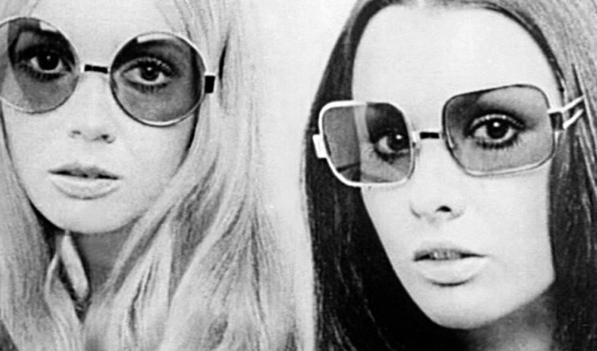 A black and white image from the 1970s of two women, blond and brunette, who stare at the camera, lips parted, with the same still, unsmiling expression. Both wear translucent oversized sunglasses.