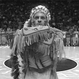 A black and white still from the film In Whose Honor? A white man in a feathered headdress, face paint and a fringed leather outfit styled after traditional Native American clothing stands on a basketball court in the center of the frame. He looks straight into the camera, unsmiling. His forearms are stacked on top of each other in front of his body. A line of basketball players stand far in the background and fans fill the bleachers.