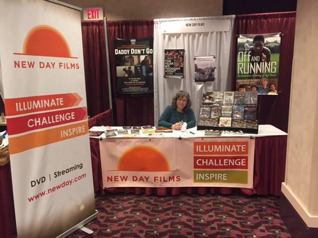 Ellen Brodsky sits at a table filled with New Day Films DVDs. Next to her, a large banner with the New Day Films' rising sun logo and "illuminate, challenge, inspire."