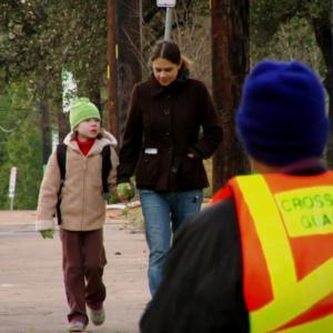 A young white mother walks hand in hand with her elementary school-aged son, approaching a school crossing guard. They're all bundled up on a chilly day.