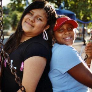 Two young girls of color stand back to back on a swing. The girl on the left is wearing a black t-shirt and hoop earrings while the girl on the right is wearing a blue t-shirt and red visor. They are both smiling.