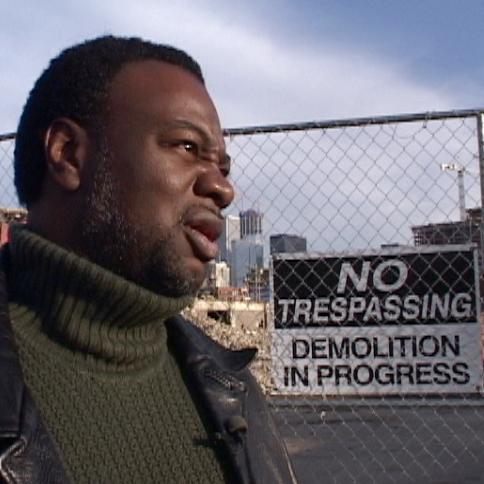 A middle-aged Black man stands outside in a turtleneck and leather jacket, frowning as he looks at something in the distance. Just past his shoulder, a high chain link fence with a sign reading "No trespassing. Demolition in progress."